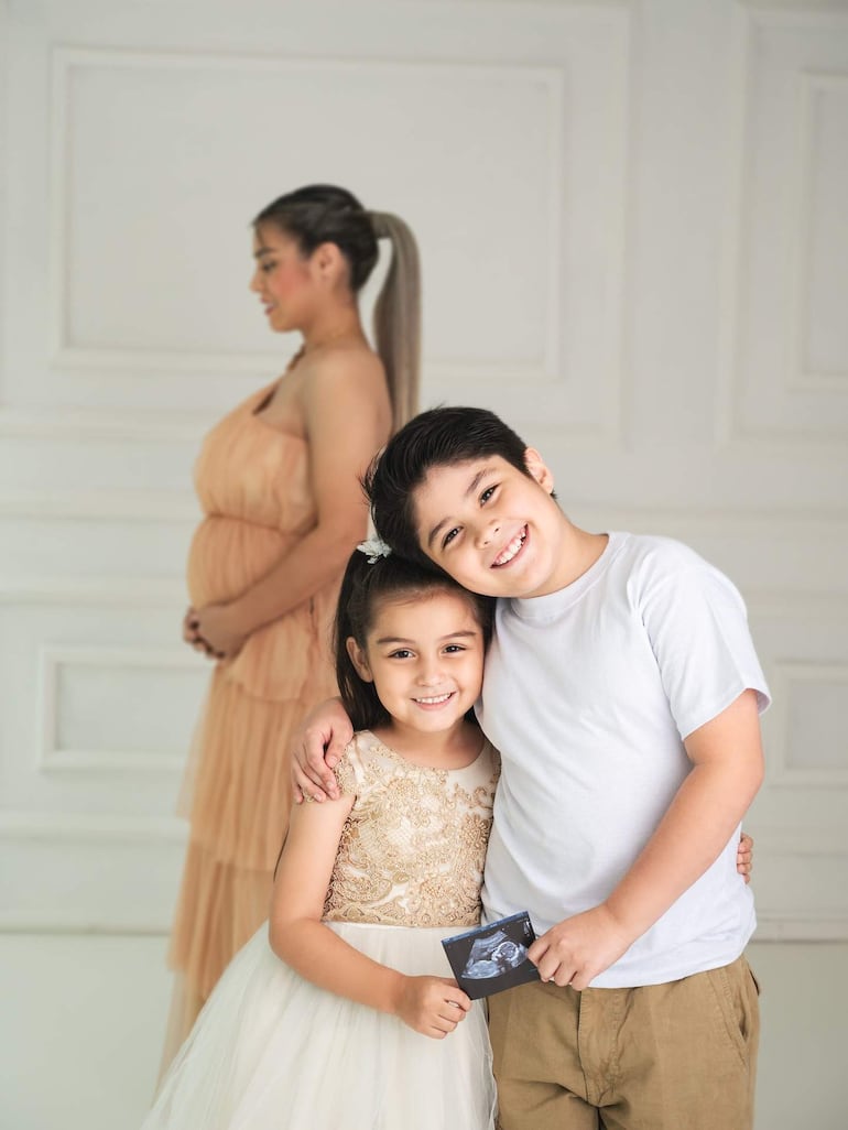 Nadia Portillo, embarazada, sonríe en vestido melocotón junto a sus hijos, un niño y una niña con vestimenta formal y expresiones felices.