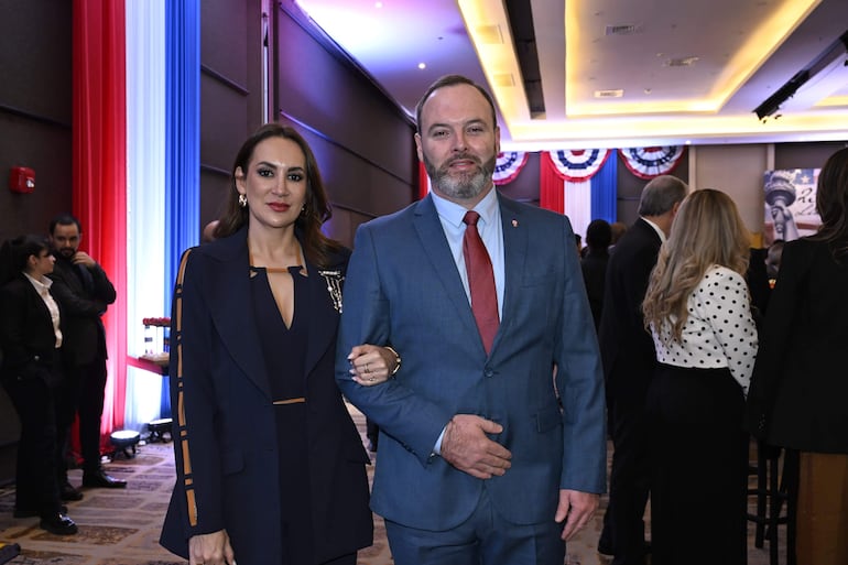Mujer con chaqueta oscura sonríe junto a hombre en traje azul y corbata roja en un ambiente decorado con colores patrios.