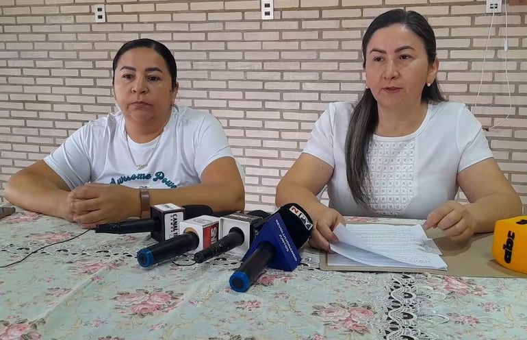 Liliana Urbieta con blusa blanca y Norma Urbieta en camiseta blanca, leyendo un comunicado ante micrófonos en ambiente de conferencia.