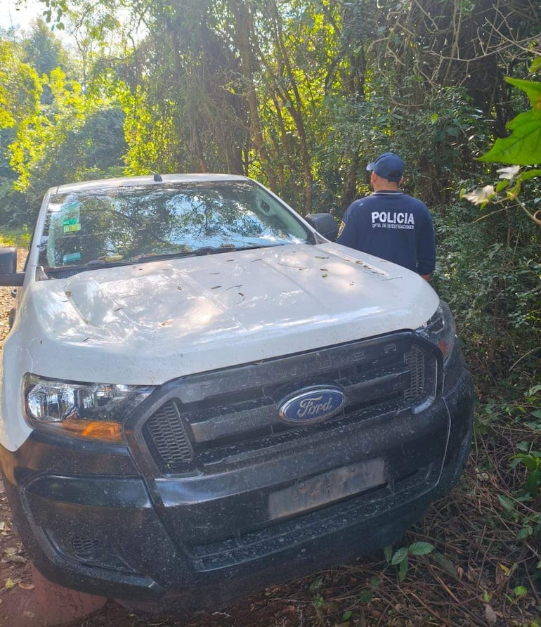 Oficial de policía de espaldas en zona boscosa, junto a una camioneta Ford blanca cubierta de hojas y suciedad.