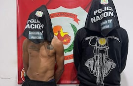 Dos personas con capuchas; una parcialmente desnuda y con tatuaje, otra con camiseta negra, frente a cartel policial.