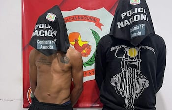 Dos personas con capuchas; una parcialmente desnuda y con tatuaje, otra con camiseta negra, frente a cartel policial.