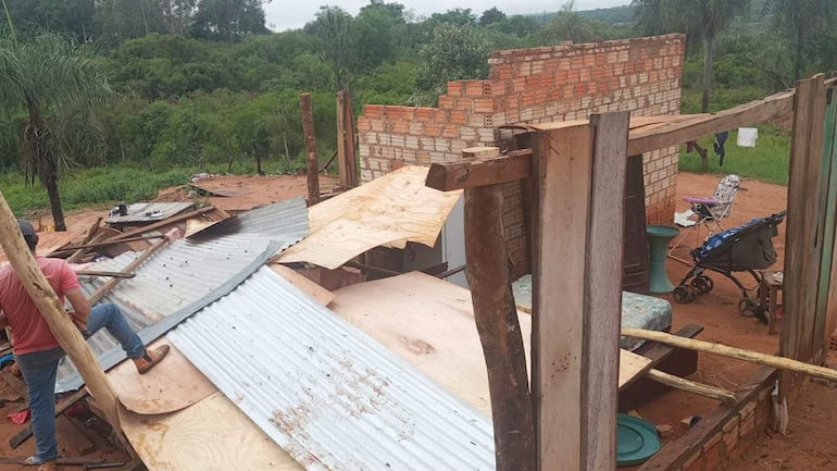 Hombre con camiseta roja trabajando en la estructura dañada, con madera y chapas esparcidas en un entorno rural.