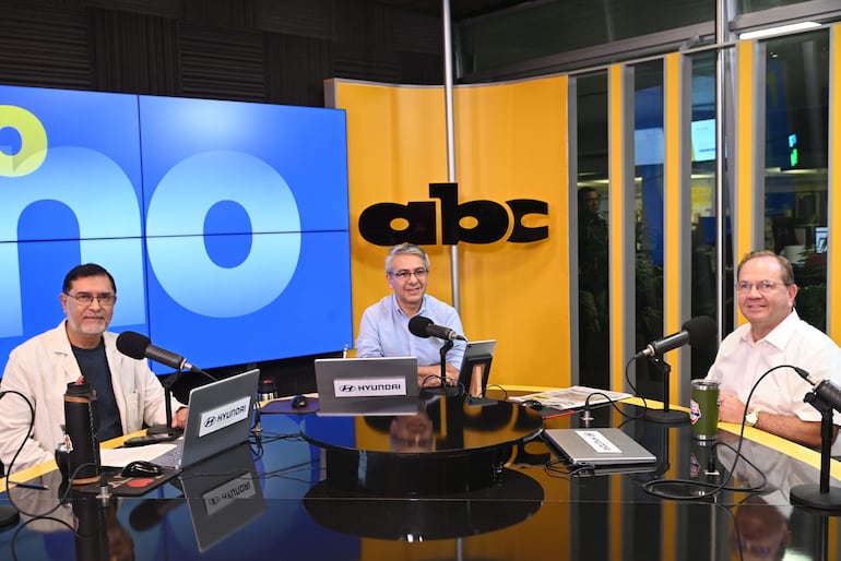 Camilo Pérez, con chaqueta clara, sonríe mientras conversa en estudio con hombre de camisa azul y otro en camisa blanca.