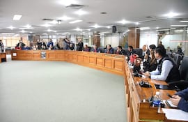 Juan José Arnold y Miguel Sosa entre asistentes en salón oficial, vestidos formalmente, expresiones serias y documentos sobre la mesa.