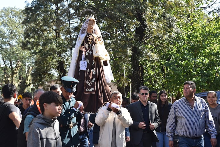 Multitud celebra al aire libre con la Virgen del Carmen, hombres con chaqueta clara y uniformes, rodeados de árboles en un día soleado.