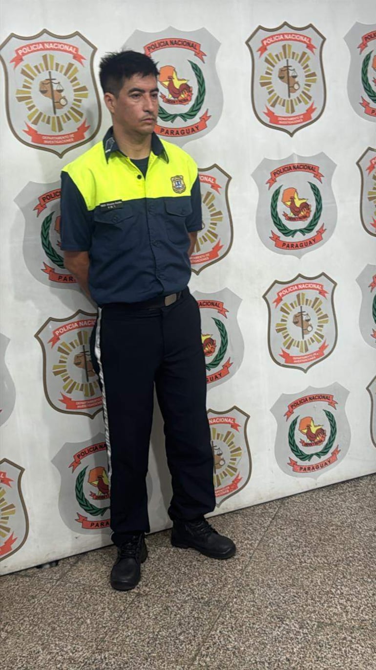 Hombre en uniforme de policía con expresión seria, frente a una pared con escudos de la Policía Nacional de Paraguay.
