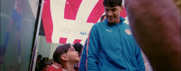 Gustavo Gómez, con chaqueta azul, sonríe mientras un niño en camiseta roja le devuelve la sonrisa en un ambiente de fútbol.