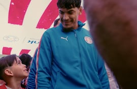 Gustavo Gómez, con chaqueta azul, sonríe mientras un niño en camiseta roja le devuelve la sonrisa en un ambiente de fútbol.