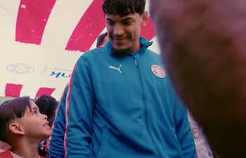 Gustavo Gómez, con chaqueta azul, sonríe mientras un niño en camiseta roja le devuelve la sonrisa en un ambiente de fútbol.