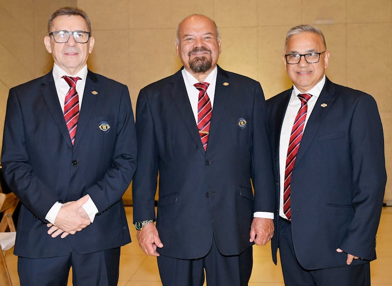 Tres hombres mayores, todos con trajes oscuros y corbatas rojas, sonríen en un ambiente formal.