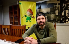Blake Webb sentado en mesa con camiseta verde y gorra negra, fondo artístico y un teléfono móvil sobre la mesa.