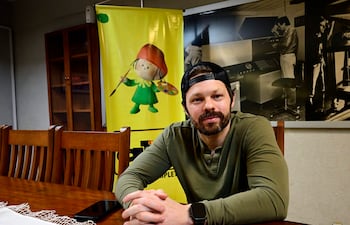 Blake Webb sentado en mesa con camiseta verde y gorra negra, fondo artístico y un teléfono móvil sobre la mesa.