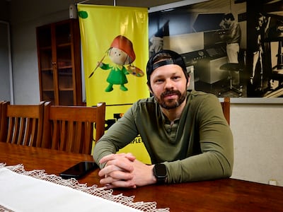 Blake Webb sentado en mesa con camiseta verde y gorra negra, fondo artístico y un teléfono móvil sobre la mesa.