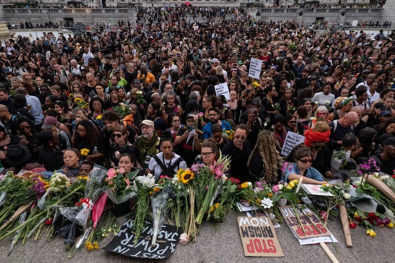 Multitud vestida de negro sostiene flores y carteles con mensajes de conmemoración, mostrando emociones de solemnidad y respeto.