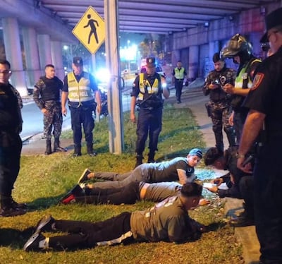 Tres hombres detenidos boca abajo, vestidos con camisetas grises, rodeados por policías en un entorno urbano con luces de emergencia.
