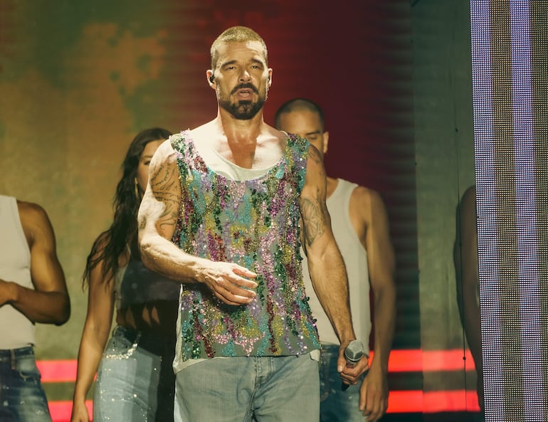 Ricky Martin con atuendo colorido y micrófono, en un escenario iluminado, acompañado de tres músicos con vestimenta sencilla.