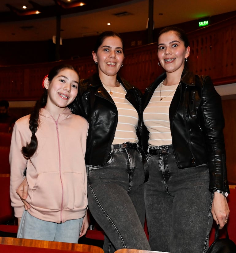 Niña en sudadera rosa sonríe junto a dos mujeres gemelas con camisetas a rayas y chaquetas de cuero en un teatro.