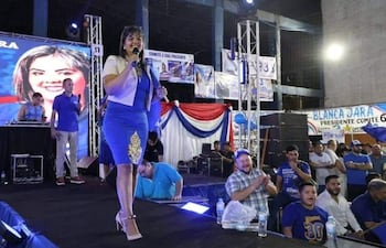 Mujer en vestido azul con dorados habla al micrófono, hombres en camisetas azules aplauden en el fondo.