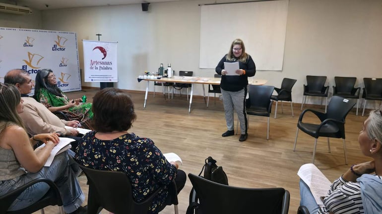Mujer de blusa negra y pantalones de cuadros leyendo ante seis personas atentas en un ambiente de taller con mesas y banners.