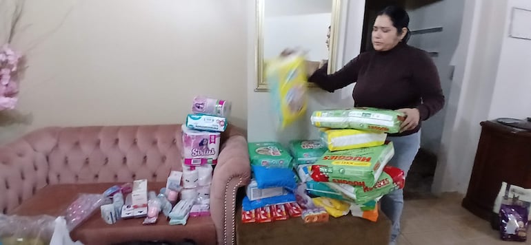 Mujer erguida sosteniendo un paquete grande, rodeada de alimentos y productos de limpieza en un salón con sofá beige.