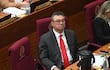 Gustavo Leite en traje oscuro y corbata roja, sentado con expresión tranquila en un salón de sesiones, rodeado de mujeres.