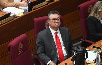 Gustavo Leite en traje oscuro y corbata roja, sentado con expresión tranquila en un salón de sesiones, rodeado de mujeres.