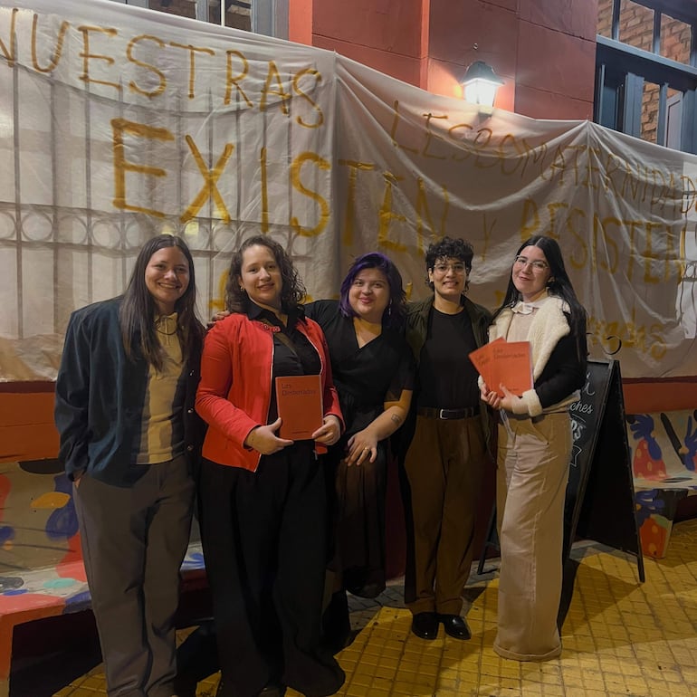 Cinco personas sonrientes sosteniendo el libro 'Las desterradas' frente a un mural colorido con un mensaje de apoyo a las lesbianas.