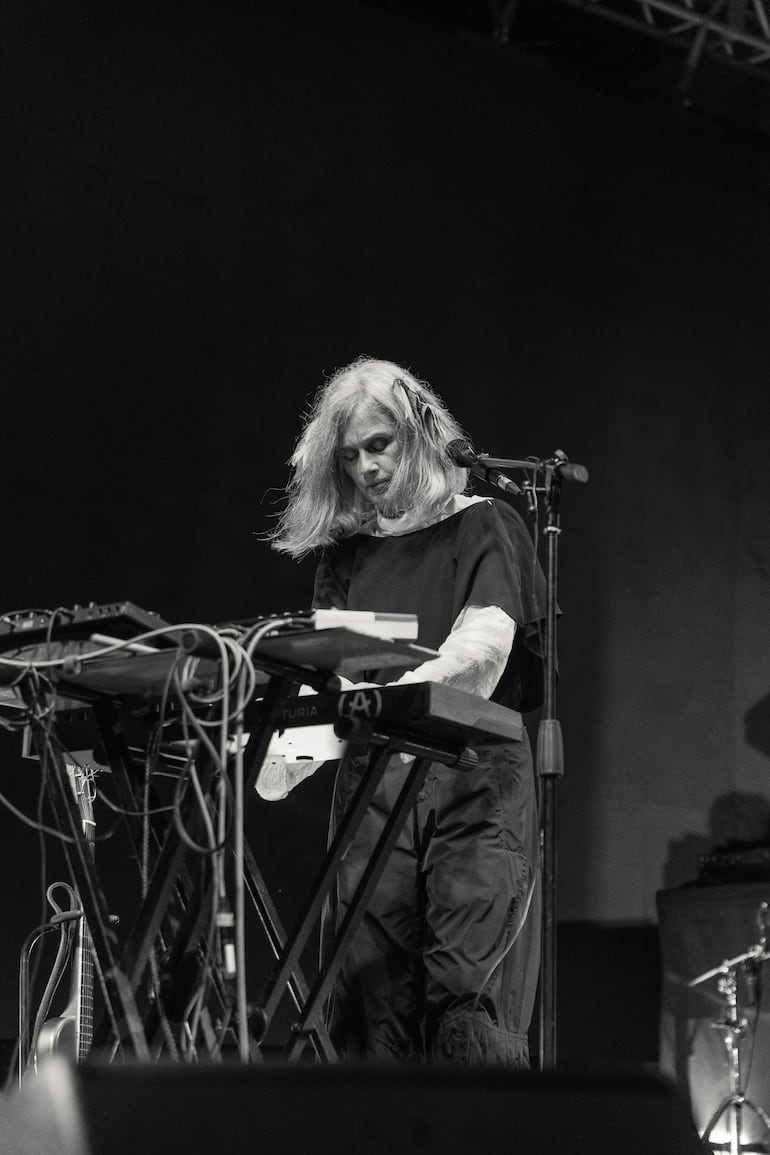 Mujer de cabello canoso, vestida de negro, tocando un teclado en un escenario oscuro con instrumentos musicales de fondo.