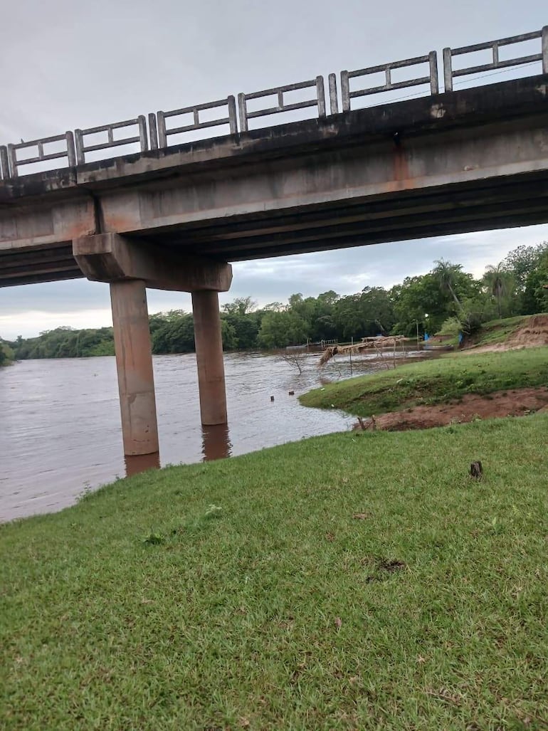 Puente elevado sobre río con césped verde en la orilla; cielo nublado sugiere inminente lluvia.