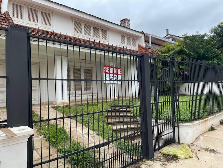 Fachada de edificio en venta con letrero rojo 'SE VENDE', rodeado de jardín descuidado y cielo nublado.