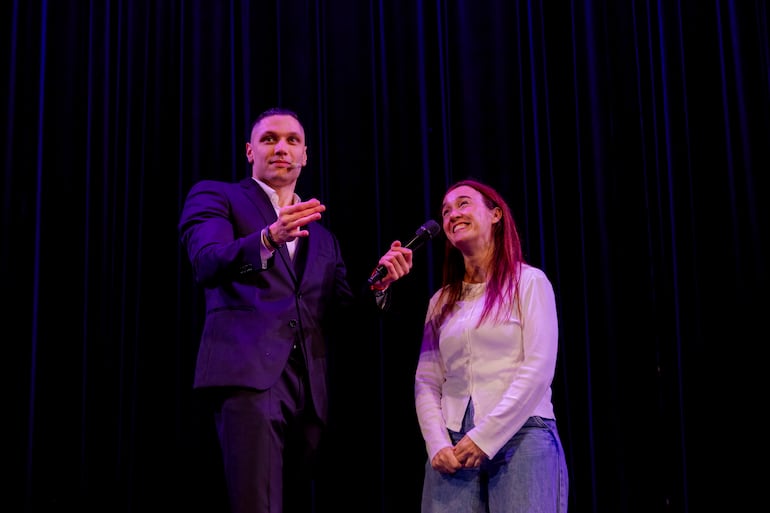 Hombre en traje oscuro presenta a mujer sonriente con blusa blanca y jeans en escenario con fondo negro.