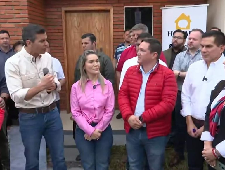 Hombre con micrófono, habla enérgicamente. A su lado, mujer con blusa rosa y otro hombre en chaqueta roja. Audiencia diversa al fondo.