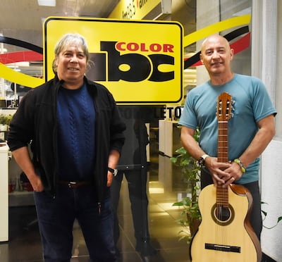 Hombre de cabello largo con suéter azul y otro con guitarra, ambos ríen frente al logotipo de ABC Color.