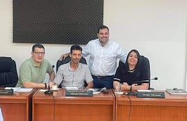 Cuatro personas en reunión: dos hombres sentados, un hombre de pie y una mujer escuchando, en un ambiente de oficina con panel acústico.