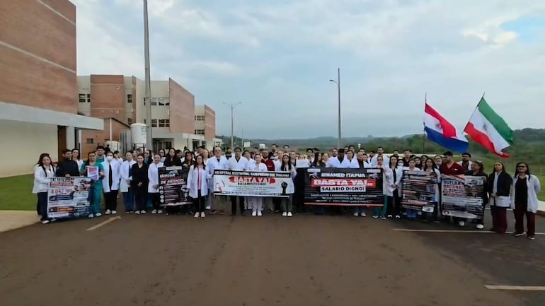 Grupo de médicos con batas blancas sostiene pancartas con mensajes de reivindicación, frente a un hospital, en una vibrante protesta.