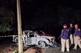 Cuatro personas, tres policías en uniforme, observan un vehículo dañado por el fuego en un ambiente rural oscuro.