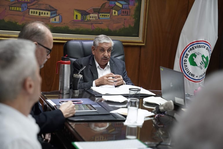 Isaías Ricardo Fretes en saco gris y camisa blanca, junto a dos hombres escuchando, en sala con bandera del IPS.