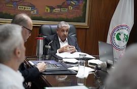 Isaías Ricardo Fretes en saco gris y camisa blanca, junto a dos hombres escuchando, en sala con bandera del IPS.