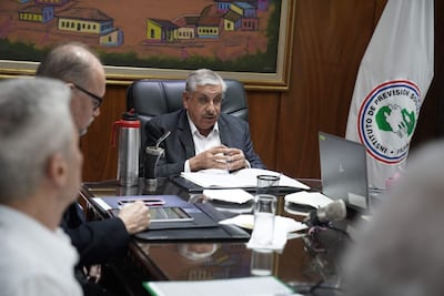Isaías Ricardo Fretes en saco gris y camisa blanca, junto a dos hombres escuchando, en sala con bandera del IPS.