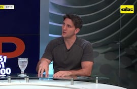 Zucolillo con camiseta gris gesticula en primer plano en set de televisión moderno, con un vaso de cristal frente a él.