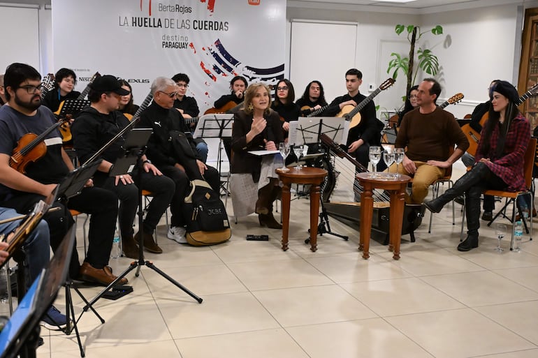 Berta Rojas en vestimenta clara habla a músicos en conferencia, rodeada de instrumentos musicales en un ambiente formal.