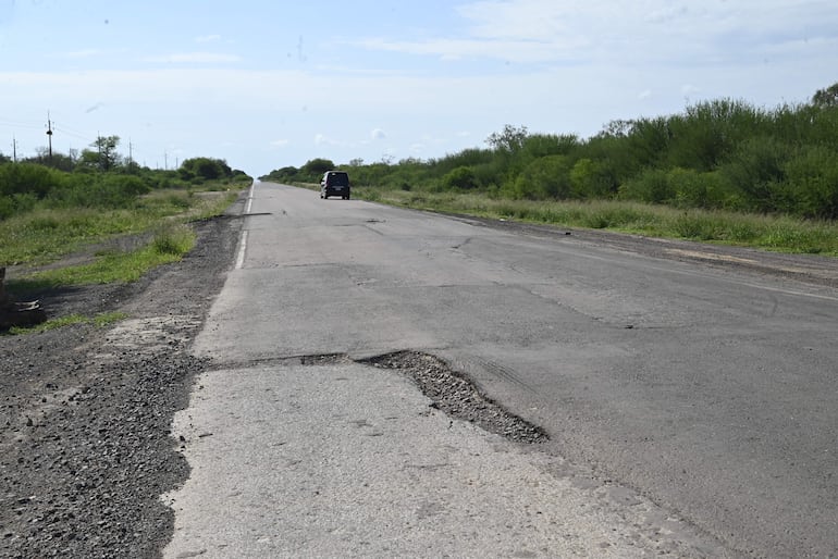 Carretera con baches y vegetación, un automóvil oscuro avanza por el camino bajo un cielo nublado.