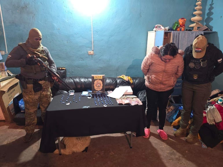 Hombre uniformado con pasamontañas sostiene un arma, mientras dos mujeres en postura de sumisión están en un ambiente interior.
