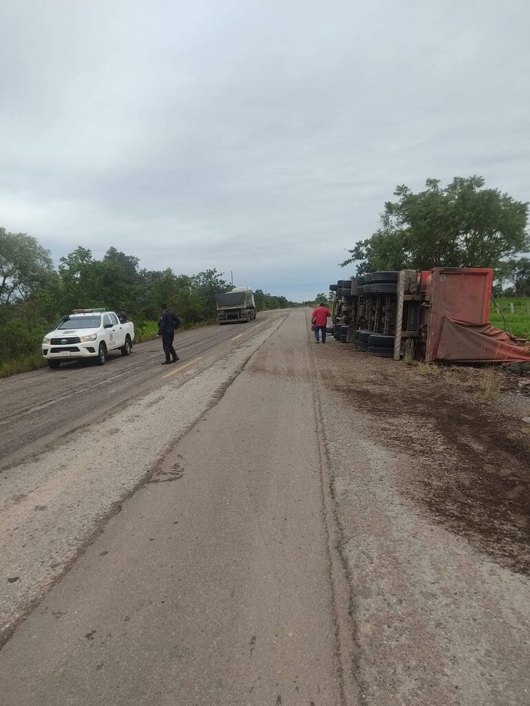 Agente en camiseta negra y otra persona en camiseta roja observan un camión grande volcado en una carretera rural con cielo nublado.