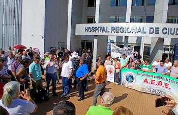 Multitud de adultos en protesta frente al hospital, destacando un hombre con altavoz y pancartas de organizaciones educativas.