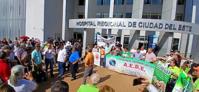 Multitud de adultos en protesta frente al hospital, destacando un hombre con altavoz y pancartas de organizaciones educativas.