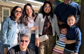 Ocho personas sonriendo en un jardín, con un hombre agachado al frente y dos niños abrazados al centro, entre árboles y una construcción.