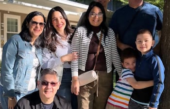 Ocho personas sonriendo en un jardín, con un hombre agachado al frente y dos niños abrazados al centro, entre árboles y una construcción.