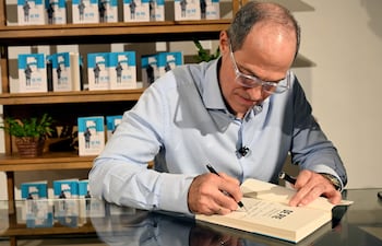 Hombre con gafas y camisa clara firma un libro, rodeado de copias del mismo en un ambiente interior.